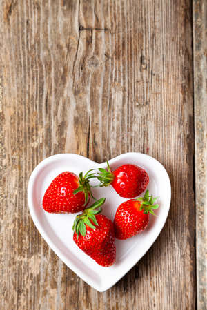 Ripe strawberries in a heart shaped plate on a wooden background. Still life with delicious berries.の写真素材
