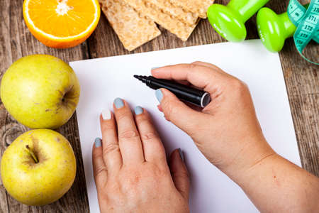 Fat woman makes a diet plan. Healthy food, dumbbells and measuring tape on a wooden background.の写真素材