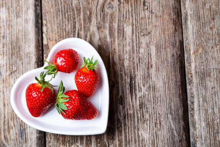 Ripe strawberries in a plate in the shape of a heart on a wooden background.の写真素材