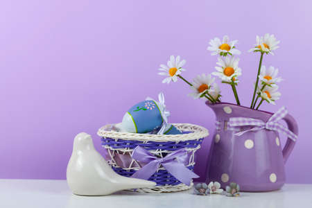 Easter eggs in a basket, a jug of flowers and a bird on a lilac background, space for text. easter...