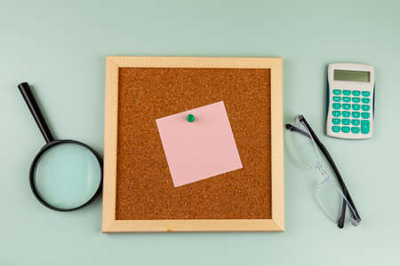 Corkboard with a note near a magnifying glass, calculator and eyeglasses on green background. flat lay compositionの写真素材