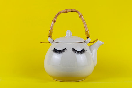 White teapot with eyes on a yellow background. Funny dishes with false eyelashes.の写真素材