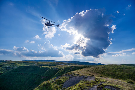 Helicopter flying over the green mountains under a blue sky with cloudsの写真素材