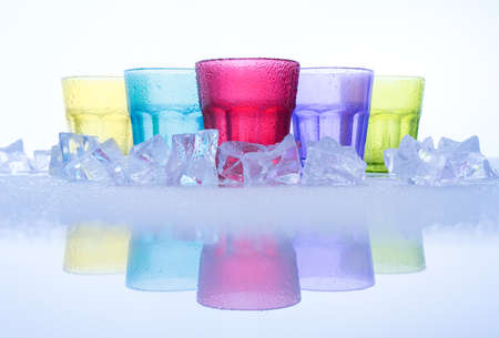 Symmetric row of multi colored of cool drinking glasses or tumbler and cube ices, with reflection on a glass table, on white backgroundの写真素材
