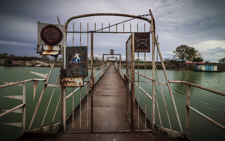 Old metal footbridge and antique gate with rusty signboard. Dark green river in a cloudy dayの写真素材