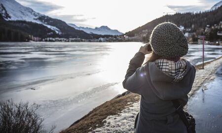 Woman takes a picture of a frozen lake with a camera during sunset in Saint Moritzの写真素材