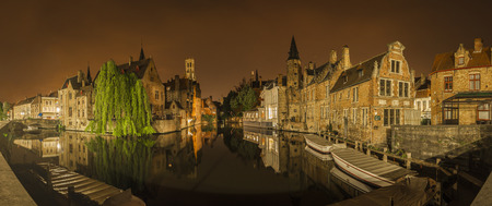 Panoramic nocturnal and iconic view of historic medieval buildings along a canal from the Rozenhoedkaai in Bruges, Belgium. Buildings, trees and boats are reflected in the waterの写真素材