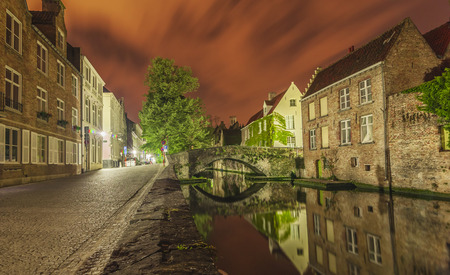 Romantic nocturnal view of a canal in Bruges. Buildings, trees and the bridge are reflected in the waterの写真素材