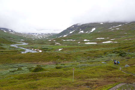 People on an hiking trail in a green valley in Sweden with a river, clouds and some snowの写真素材