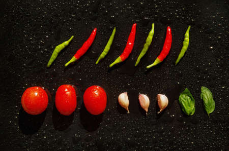 Composition of spicy, green and red chillies, two basil leaves, garlic and red tomatoes on a black background.の写真素材