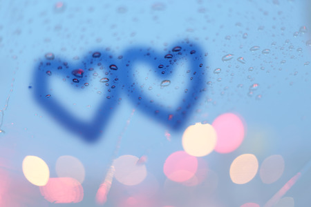 Rain drops and two hearts write on window with light bokeh, rainy season abstract background.
