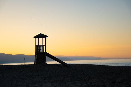 Lifeguard tower on beach at warm sunset glow, orange sky and mountains. Silhouette wooden lifeguard tower by sea. Nature, tranquility, and coastal safety landscape conceptの写真素材
