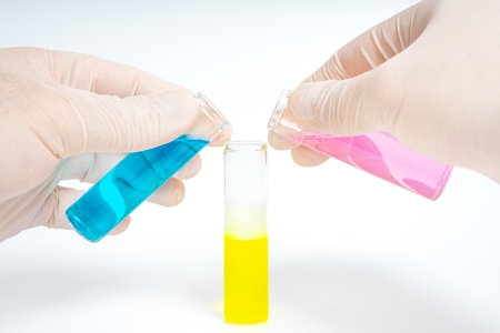 Hands holding test tubes with blue and pink liquid pouring into a test tube with yellow liquid isolated on white backgroundの写真素材