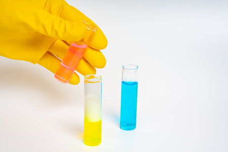Yellow gloved hands holding glass test tubes with blue, pink, and yellow liquids on isolated white...