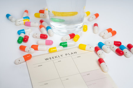 Colorful pills and water glass on weekly plan schedule sheet isolated white background. Medicine capsules. Pills and weekly planner. Health, wellness, routine, organizationの写真素材