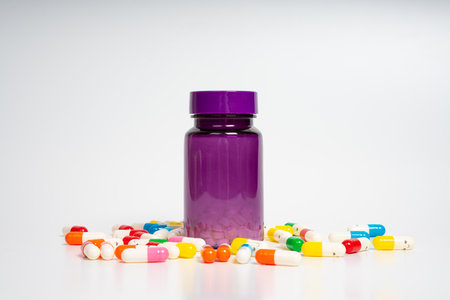 Purple pill bottle surrounded by colorful capsules on isolated white background. Closeup of medicine bottle with scattered pills. Health, medication and treatment conceptの写真素材