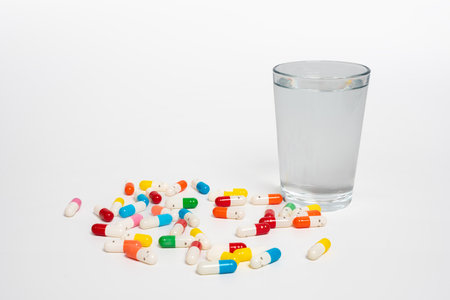 Scattered colorful capsules with a glass of water on isolated white background. Medicine and hydration with pills and water. Health, treatment and daily care conceptの写真素材