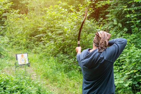 Man with bow and arrow aiming at archery target in forest, aiming an arrow at a target, back view,...