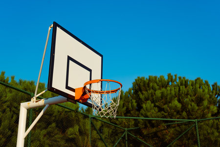 Basketball hoop with orange rim and net, green trees, blue sky. Basketball goal on fence. Sports, outdoor activity and recreation conceptの写真素材