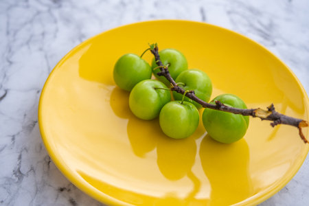Fresh ripe green plums on yellow plate with branch on marble background surfaceの写真素材