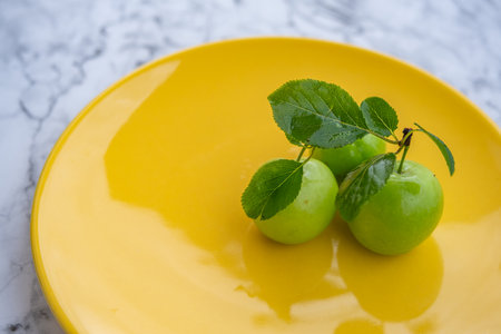 Fresh ripe green plums on yellow plate with branch on marble background surfaceの写真素材