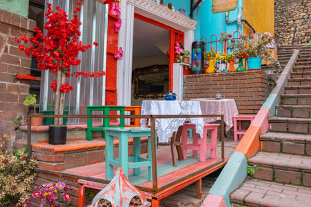 Colorful outdoor cafe table and chairs with vintage wooden stools and flowers on cozy brick terrace. Charming rustic street cafe with small wooden tables, bright colors, stairs and floral decorの写真素材