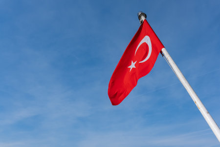 Red Turkish flag with white star and crescent flying on flagpole, sunny day background. Waving in the wind against bright blue sky, national symbol of Turkey. Copy spaceの写真素材