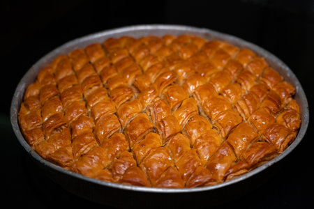 Large, round metal tray filled with freshly baked, glossy, diamond-cut traditional Turkish or Middle Eastern baklava. Detail of traditional handmade baklava. Pastry dessert isolated on dark backgroundの写真素材