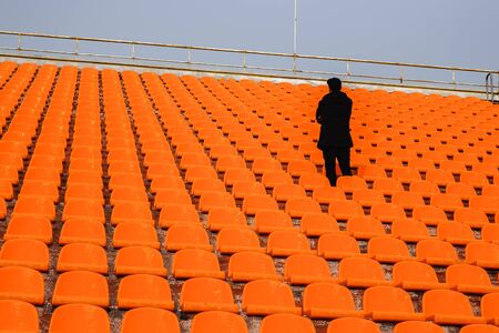 Young man stands in stadium from orange chairs.