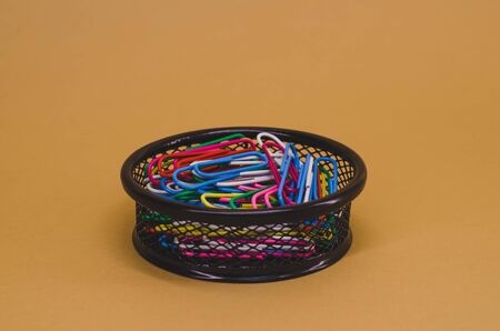 pile of multicolored paper clips lying in a black basket against a brown background.