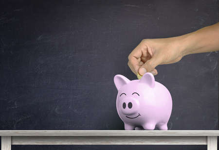human hand dropping a coin into white piggy bank on wood shelf with against chalkboard backgroundの写真素材