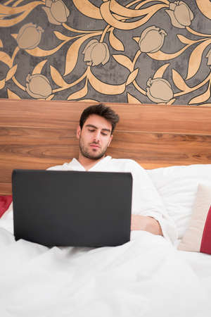 Attractive young man in white robe in his hotel room surfing on his laptopの写真素材