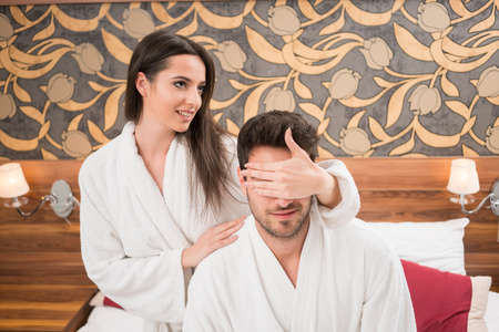 Beautiful happy young couple enjoying in bed after spa. Couple in white robes relaxing, while woman is covering mans eyes with her armの写真素材