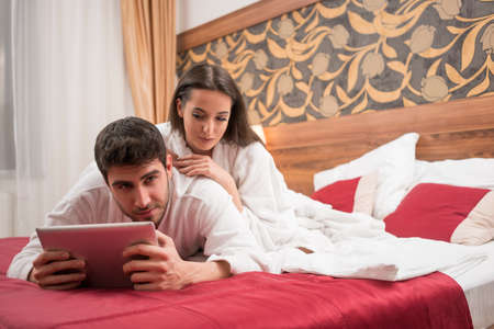 Beautiful happy young couple enjoying in bed after spa. Couple in white robes relaxing. Man using tablet while having conversation with womanの写真素材