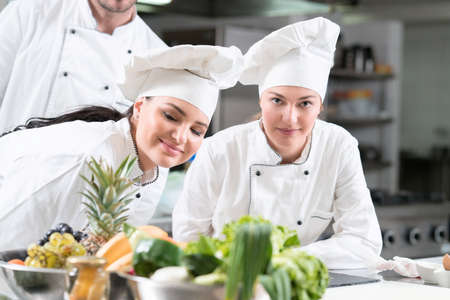 Two glad female young cooks wearing uniform working on kitchen. Preparing delicious meal.の写真素材