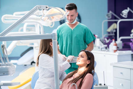 people, medicine, stomatology and health care concept - happy female dentist with mirror checking patient girl teeth up at dental clinic office while male assistant is waiting behind.の写真素材
