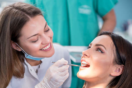 people, medicine, stomatology and health care concept - happy female dentist with mirror checking patient girl teeth up at dental clinic office while male assistant is waiting behind.の写真素材