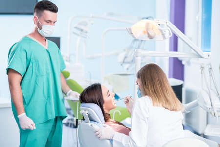 people, medicine, stomatology and health care concept - happy female dentist with mirror checking patient girl teeth up at dental clinic office while male assistant is waiting behind.の写真素材