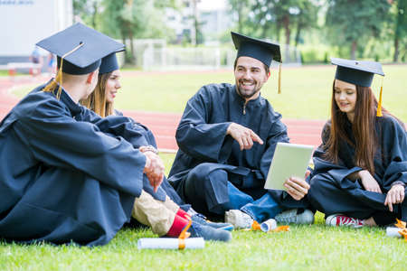 Group of Diverse International Graduating Students Celebrating, sitting on a grass, relaxing and having fun. Happy young students relaxing on lawn in graduation clothesの写真素材
