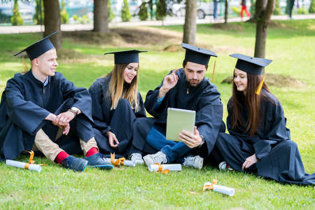 Group of Diverse International Graduating Students Celebrating, sitting on a grass, relaxing and having fun. Happy young students relaxing on lawn in graduation clothesの写真素材