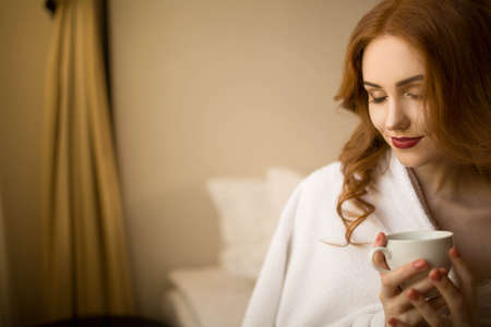 Red-haired girl in a white robe with a cup of coffee sitting on a bedの写真素材