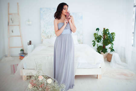 Cute girl with makeup in a blue evening dress standing beside the bed. Beautiful cozy interior in the backgroundの写真素材