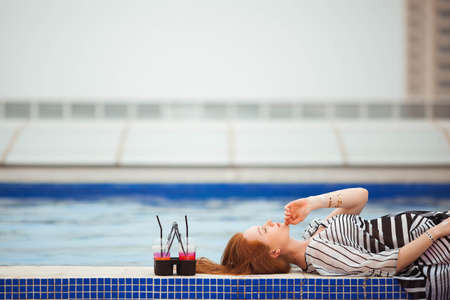 Beautiful red-haired girl in a striped pareo lying by the pool. Next are the cocktails.の写真素材