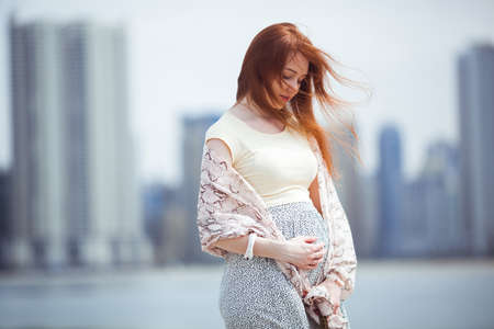 Redheaded pregnant woman touching her belly with skyscrapers in the background. Summerの写真素材