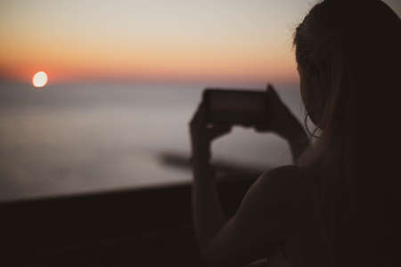 Beautiful blonde girl shoots sunset on her phone. Close up.の写真素材