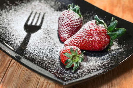 three strawberries sprinkled with sugar on a black plate and wooden baseの写真素材
