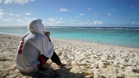 A man with a deep-thinking head and a towel over his head looking at the blue sea.の写真素材