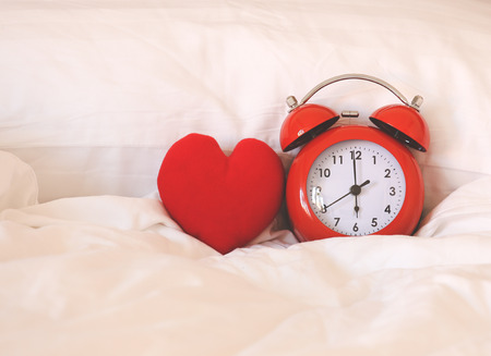 Red alarm Clock and heart shape on white bed sheet against the pillow, sleep well concept