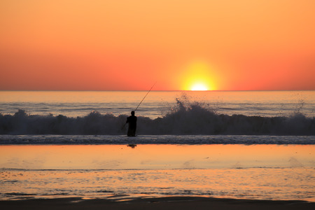 Silhouette of fisherman hloding fishing pole on the beach at sunsetの写真素材