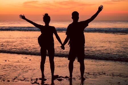 Silhouettes of a couple at sunset, arms raised up showing happiness and healthy lifestyle against a colourful sky of clouds background.の写真素材